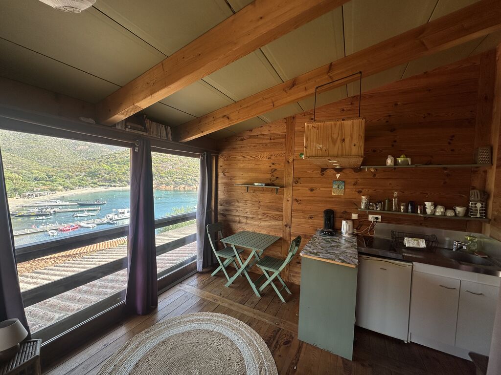 Coin cuisine et table bistrot du studio, avec le port de Girolata derrière la baie vitrée